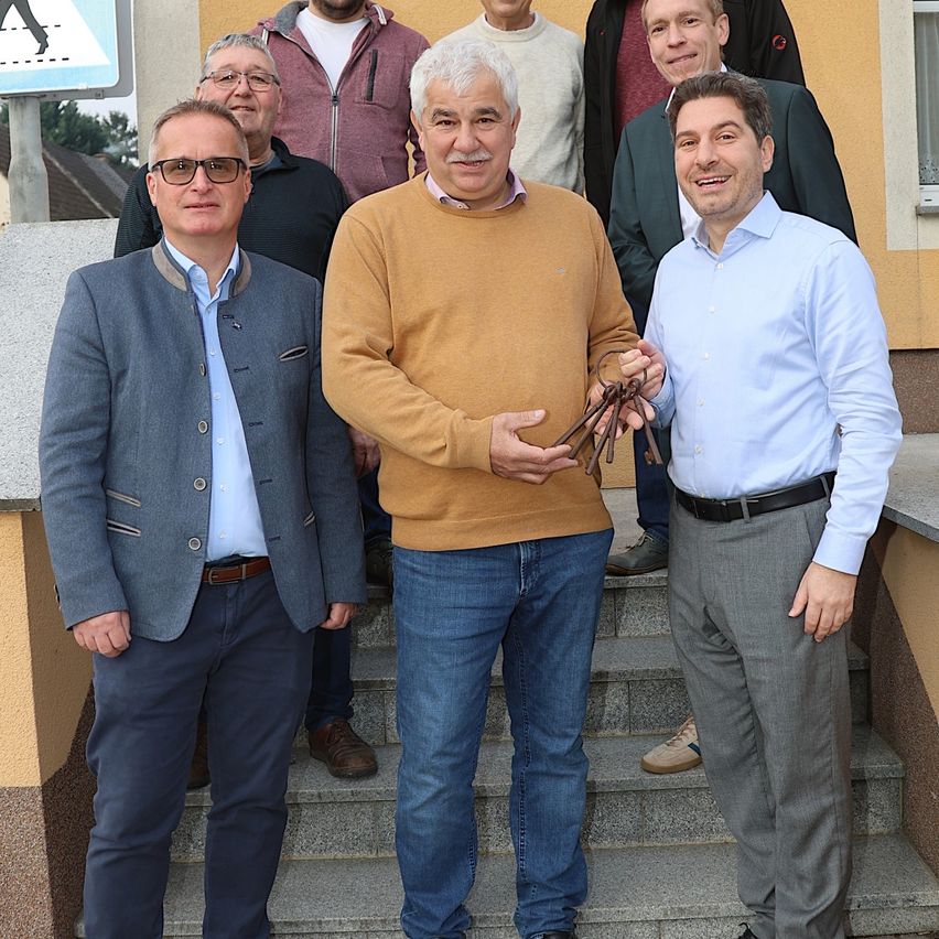 Eine Gruppe von Männern steht vor einem Gebäude auf Stufen. Sie tragen alle formelle Kleidung. Der Mann in der Mitte hält einen Schlüssel.