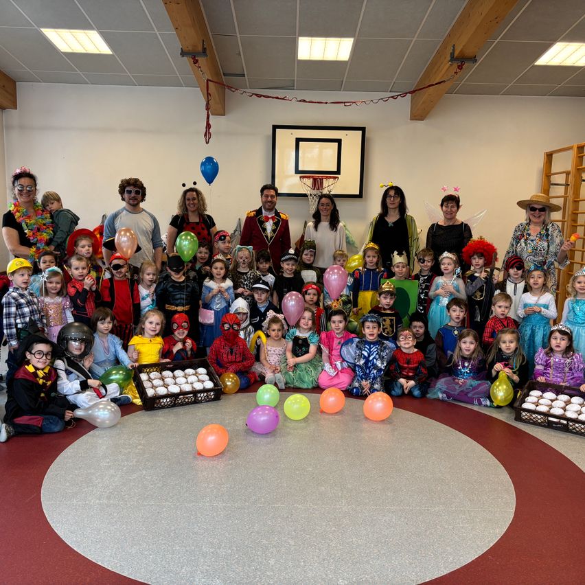 Eine Gruppe von Menschen posiert für ein Foto in einem Raum, einige in Kostümen und mit Luftballons. An der Wand befindet sich ein Basketballkorb und auf dem Boden stehen Körbe mit Cupcakes.