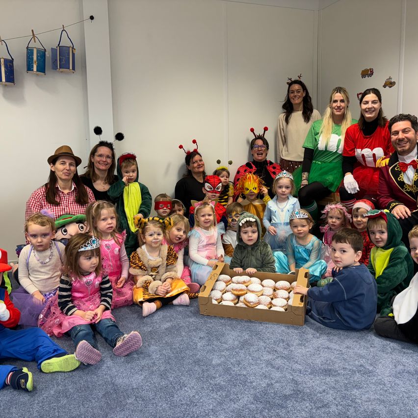 Eine Gruppe von Kindern und Erwachsenen in verschiedenen Kostümen sitzt und steht um eine Box mit Donuts auf dem Boden. Einige tragen Masken. Eine Wand im Hintergrund hat Dekorationen und Text.