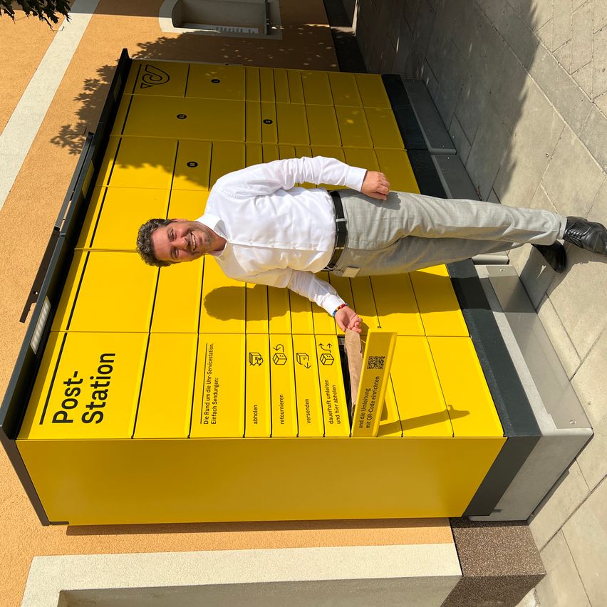 Ein Mann in einem weißen Hemd und grauen Hosen liegt auf einem gelben Post Station Schließfach, lächelt und hält eine Box.