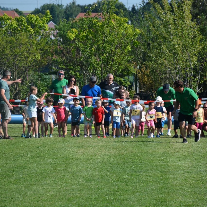 Kinder stehen für ein Rennen auf einem grünen Feld an. Sie haben Nummern auf ihren Trikots. Erwachsene stehen hinter ihnen, einige tragen Hüte. Bäume und Gebäude sind im Hintergrund zu sehen.