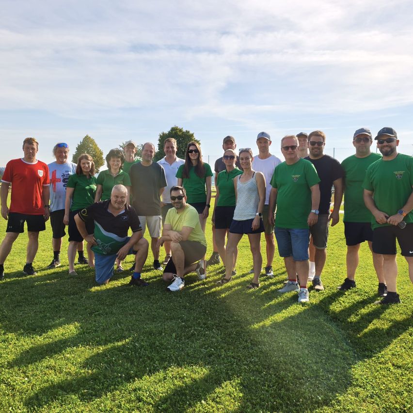 Eine Gruppe von Menschen in grünen Shirts posiert für ein Foto auf einem Rasenfeld mit Bäumen im Hintergrund und einem teilweise bewölkten Himmel.
