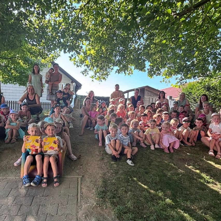 A group of people, including adults and children, are gathered on a grassy area. They are posing for a photo with a house in the background. Some are sitting on a bench while others are standing.