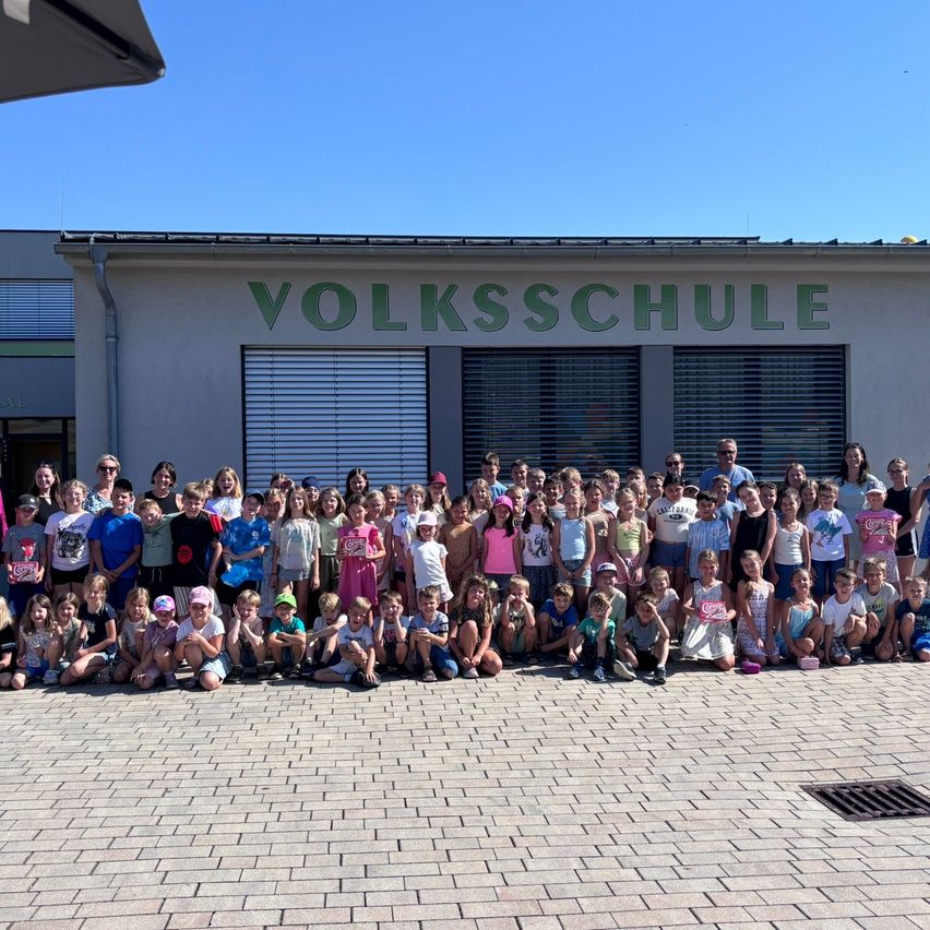 A large group of children and adults are gathered in front of a building with the sign Volksschule. They are all smiling and posing for a picture.