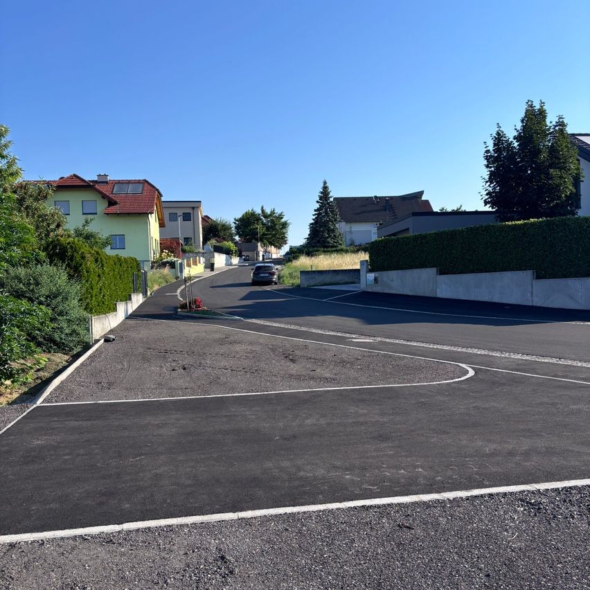 A residential street with houses, a car parked, and greenery along the edges. The road is empty, with white markings.