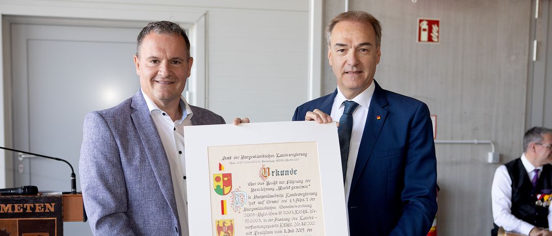 Zwei Männer in Anzügen und Krawatten halten ein gerahmtes Zertifikat, lächeln, einer hält einen roten Wachssiegel. Das Zertifikat trägt den Titel 'Urkunde' und hat Text und Logos auf Deutsch.