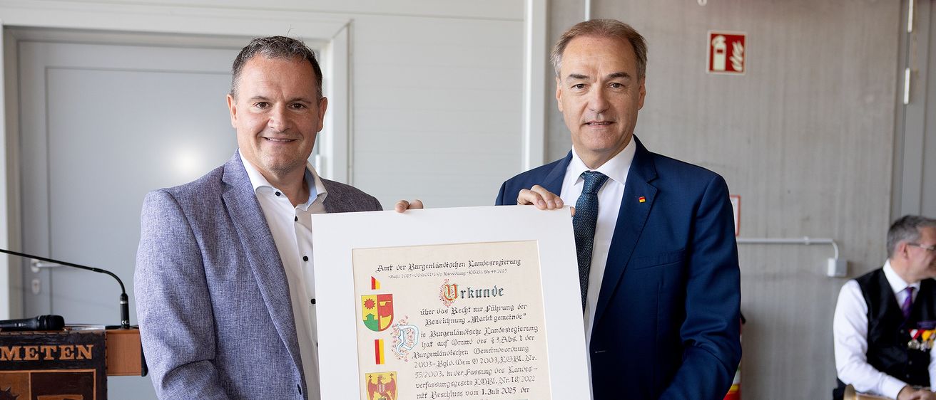 Zwei Männer in Anzügen und Krawatten halten ein gerahmtes Zertifikat, lächeln, einer hält einen roten Wachssiegel. Das Zertifikat trägt den Titel 'Urkunde' und hat Text und Logos auf Deutsch.