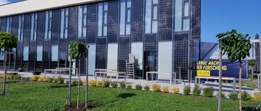 Ein modernes Gebäude mit Solarmodulen an der Fassade, umgeben von Grünpflanzen und Tischen draußen. Der Himmel ist klar mit einigen Wolken.