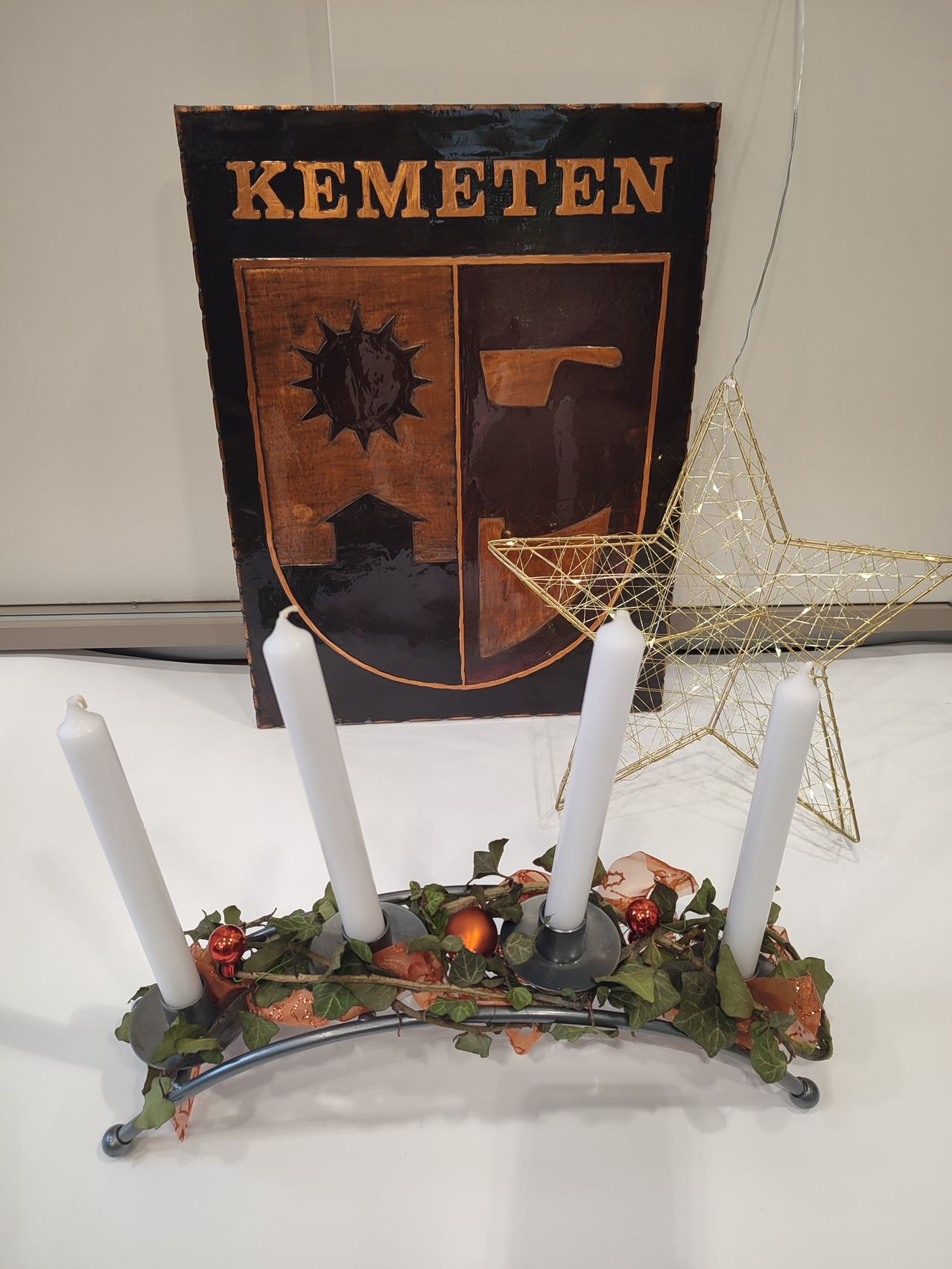 Ein Holzschild mit dem Wort 'KEMET' und einem Wappendesign steht hinter vier Kerzen und einem Stern. Die Kerzen sind in einer Reihe auf einem Ständer angeordnet.
