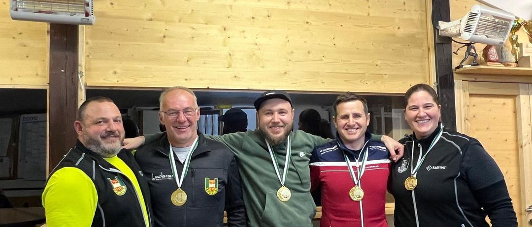 Fünf Personen mit Medaillen stehen in einer Reihe. Zwei Frauen und drei Männer, alle lächelnd, tragen Sportjacken. Die hölzerne Wand dahinter verleiht der Szene eine rustikale Note.