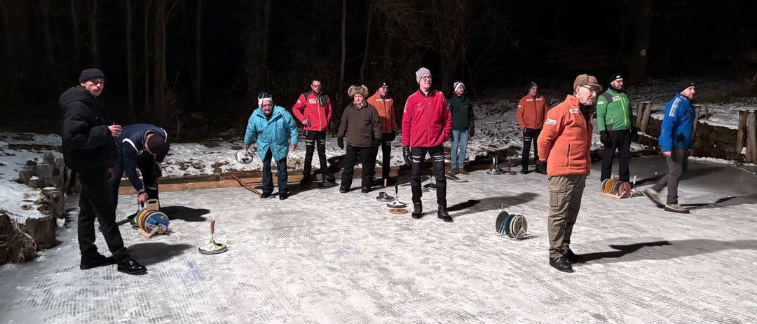 Mehrere Menschen in Winterkleidung stehen auf einer Eisbahn in einem Waldgebiet und bereiten sich auf ein Curling-Spiel vor.