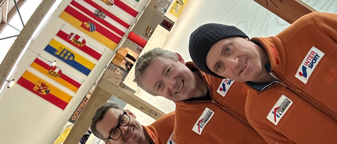 Vier Männer in orangefarbenen Overalls stehen eng beieinander und lächeln für ein Foto. Sie befinden sich in einer Sporteinrichtung mit Fahnen an der Wand.