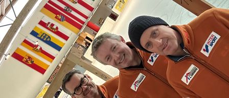 Vier Männer in orangefarbenen Overalls stehen eng beieinander und lächeln für ein Foto. Sie befinden sich in einer Sporteinrichtung mit Fahnen an der Wand.