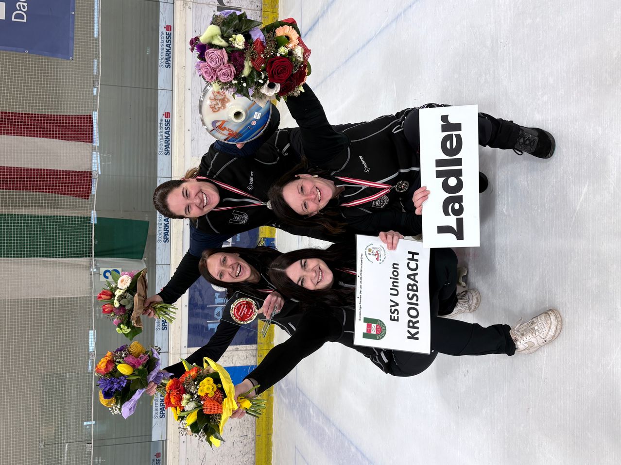Drei Frauen posieren auf einer Eisbahn, halten Blumen und Medaillen, wobei eine ein Schild mit 'Ladler' und eine andere mit 'ESV Union Kroisbach' hält.