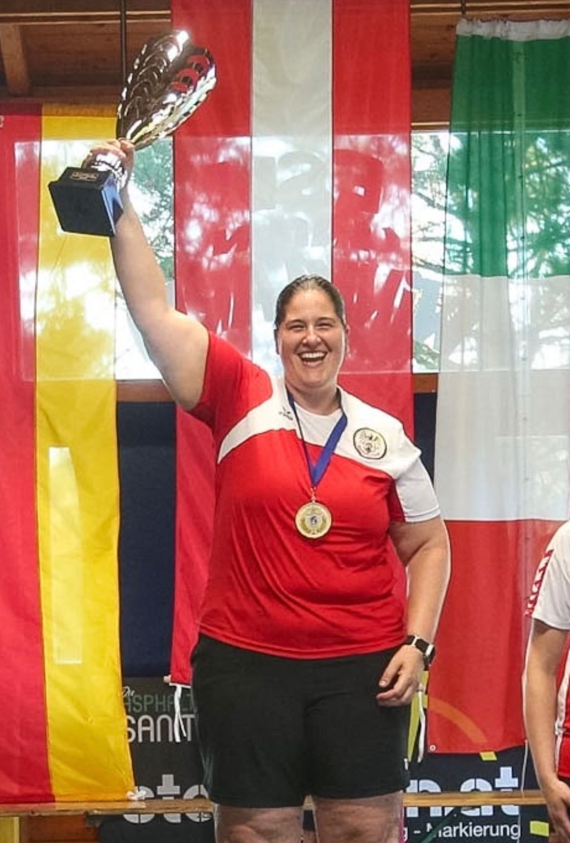 Eine Frau steht auf einem Podium mit einer Goldmedaille um den Hals, lächelt und hält eine Trophäe. Sie ist von Flaggen verschiedener Länder umgeben.