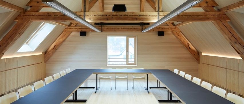 Ein Konferenzraum mit Holzbalken, einem langen Tisch und Stühlen. Ein Fenster an der Wand lässt Licht herein. Zwei Lampen hängen von der Decke.
