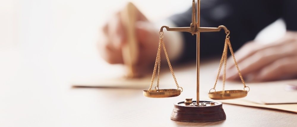 Eine goldene Waage mit Ketten auf einem Holzsockel, mit einer Hand, die ein Siegel auf einem Schreibtisch hält. Der Hintergrund ist verschwommen.