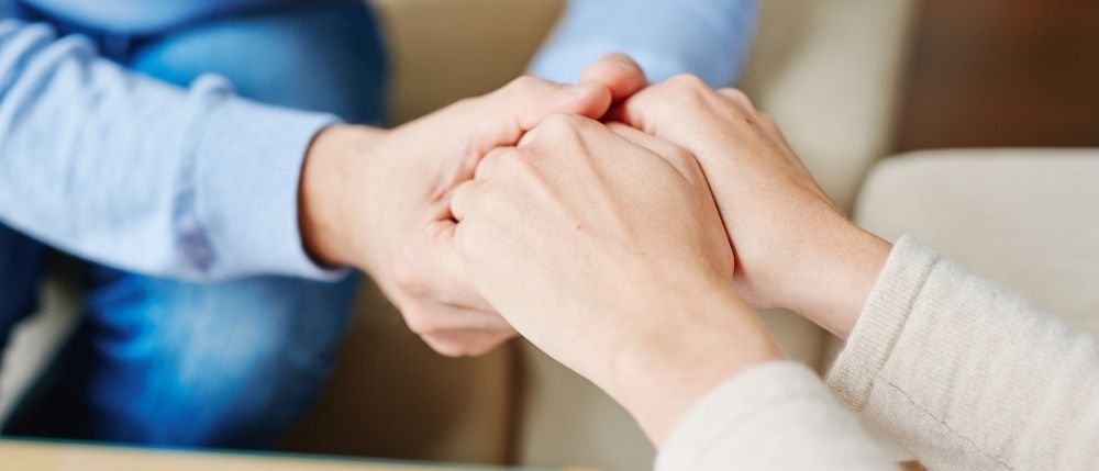 Zwei Menschen halten Händchen und zeigen einen tröstlichen Moment, einer mit einem blauen Shirt, der andere mit einem weißen Shirt.