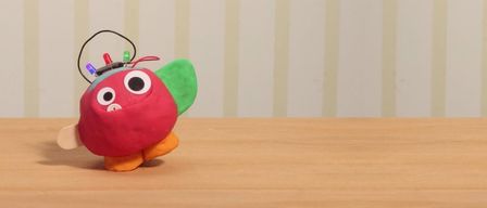 Ein rotes rundes Spielzeug mit großen Augen und einer kleinen Antenne sitzt auf einem Holztisch. Es hat grüne Flügel und orange Füße.