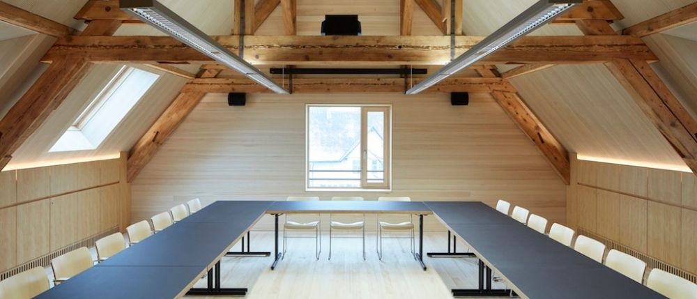 Ein leerer Konferenzraum mit langen Tischen, weißen Stühlen, Holzwänden und zwei Fenstern mit Lautsprechern darüber.