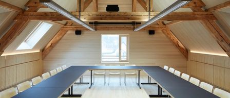 Ein leerer Konferenzraum mit langen Tischen, weißen Stühlen, Holzwänden und zwei Fenstern mit Lautsprechern darüber.
