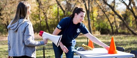 A woman in a blue shirt holds a megaphone, reaching towards a table with orange cones and gloves. Another woman looks on from the side.