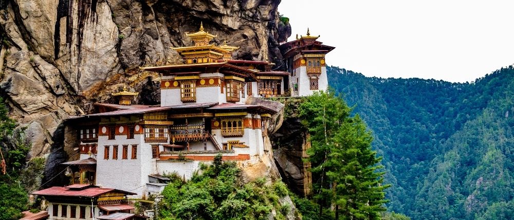 A traditional Bhutanese monastery perched on a cliff with a lush green mountain backdrop.