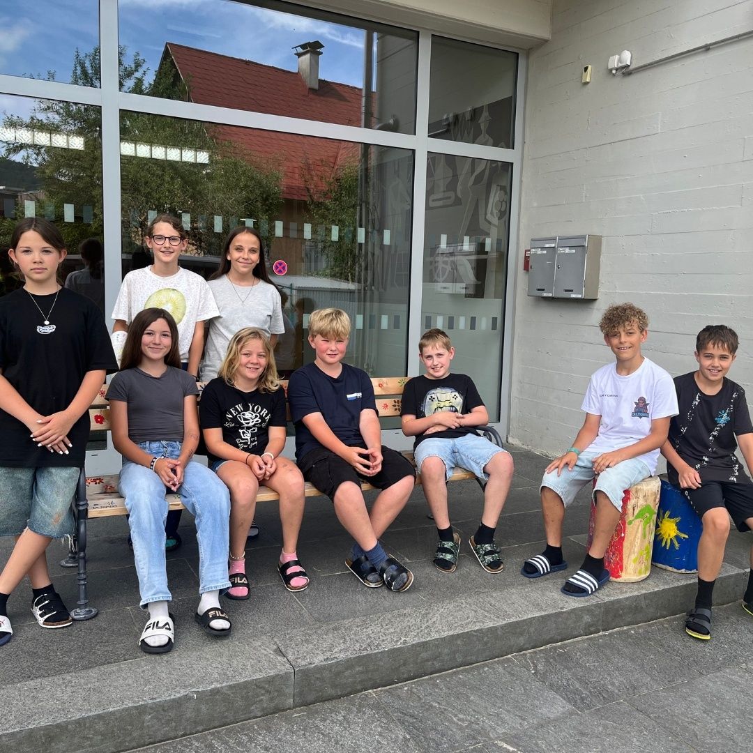 Eine Gruppe von Kindern posiert für ein Foto vor einem Gebäude mit Glaswänden, lächelt und sitzt auf einer Bank. Sie tragen Freizeitkleidung.