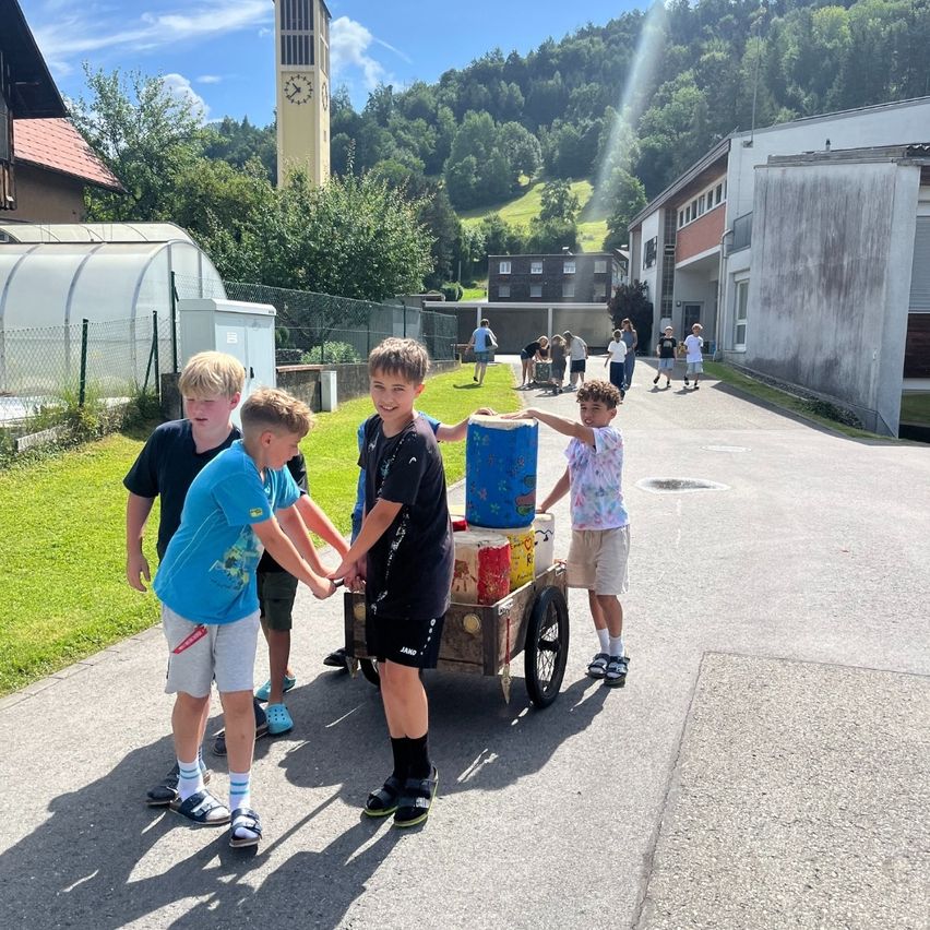 Eine Gruppe von Jungen schiebt einen Wagen mit Eimern. Sie stehen auf einer Straße mit Gebäuden und Bäumen im Hintergrund.
