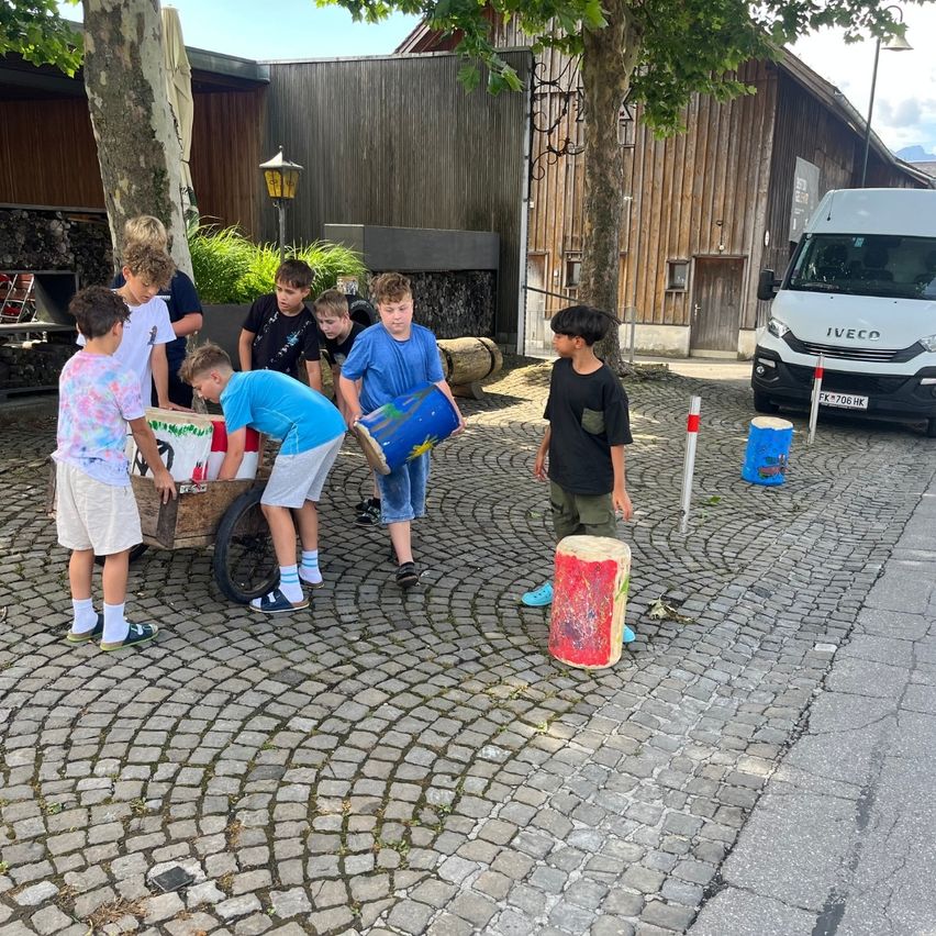 Eine Gruppe von Kindern spielt auf einer Kopfsteinpflasterstraße mit einem Wagen und Eimern. Ein weißer Van ist in der Nähe geparkt.