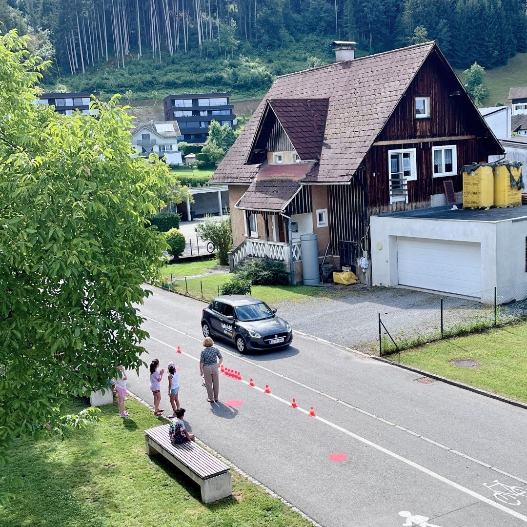 Auf einem Hügel steht ein Haus mit Garage in der Nähe einer Straße, wo ein geparktes Auto und mehrere Personen bei roten Kegeln stehen. In der Nähe gibt es eine Bank, Bäume und Gebäude.