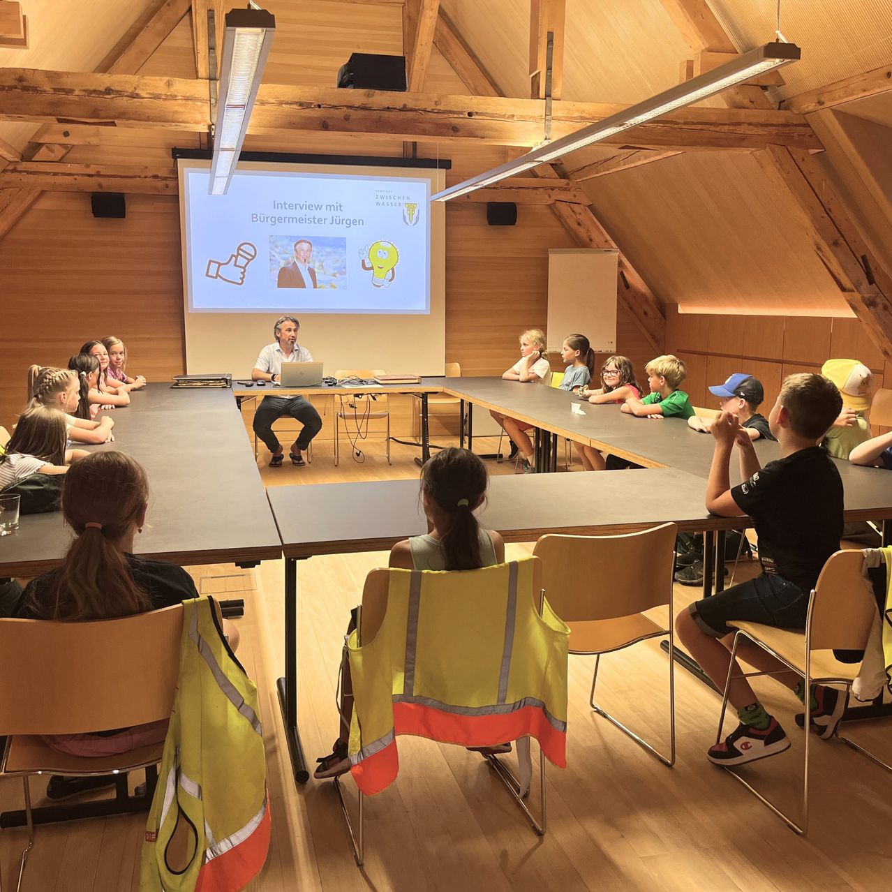 Eine Gruppe von Kindern sitzt um einen Tisch herum in einem Raum und sieht sich eine Präsentation auf einer Projektionsleinwand an. Ein Mann spricht in ein Mikrofon.