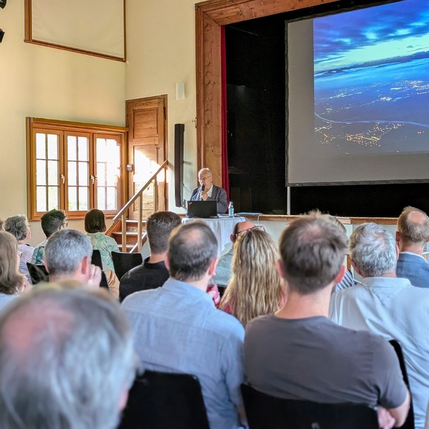 Ein Mann spricht an einem Rednerpult mit einer Projektionsleinwand hinter ihm, die eine nächtliche Stadtlandschaft zeigt. Eine Gruppe von Menschen sitzt aufmerksam davor.