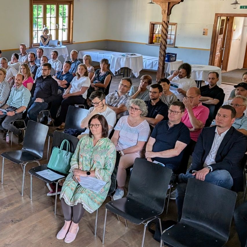 Eine Gruppe von Menschen sitzt in einem Konferenzraum, eine Frau in einem grünen Kleid hält Papiere. Einige tragen Brillen, andere haben Taschen auf dem Schoß. Tische mit weißen Tüchern sind im Hintergrund aufgestellt, und eine Tür führt in einen anderen Raum.