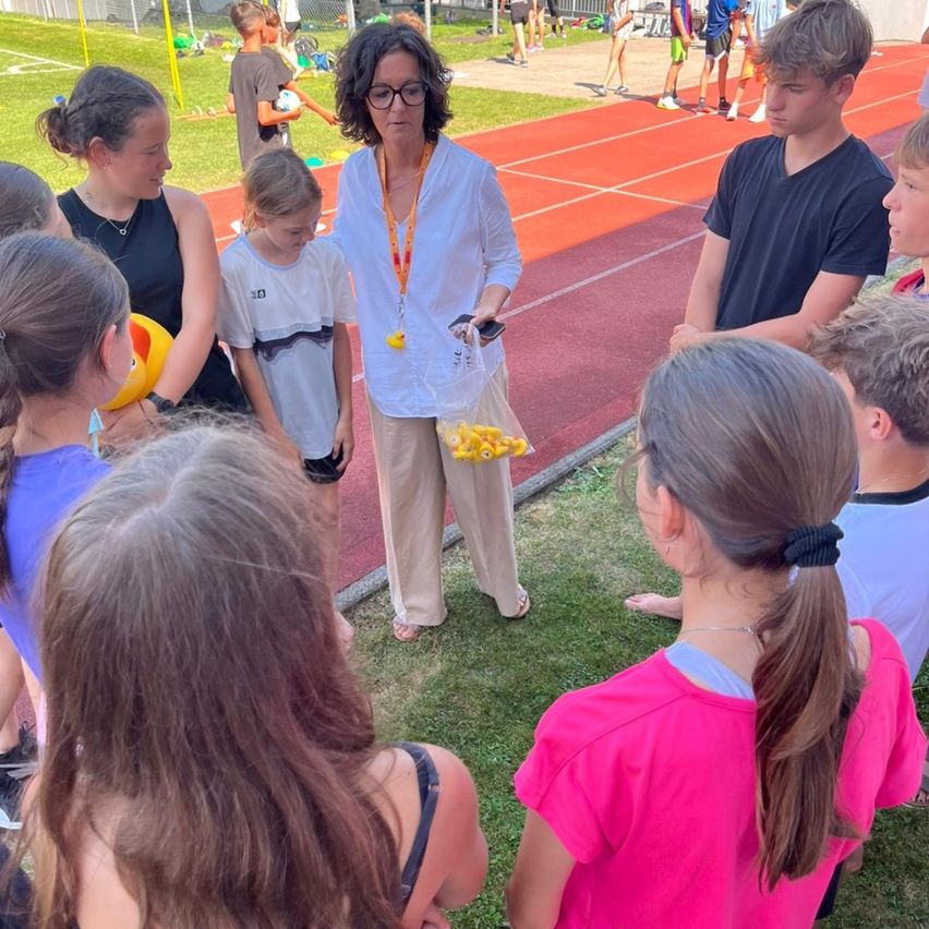 Eine Frau in einem weißen Hemd steht mit Kindern auf einer Laufbahn. Die Kinder schauen zu ihr, und sie hält eine Tasche. Andere Kinder spielen im Hintergrund.