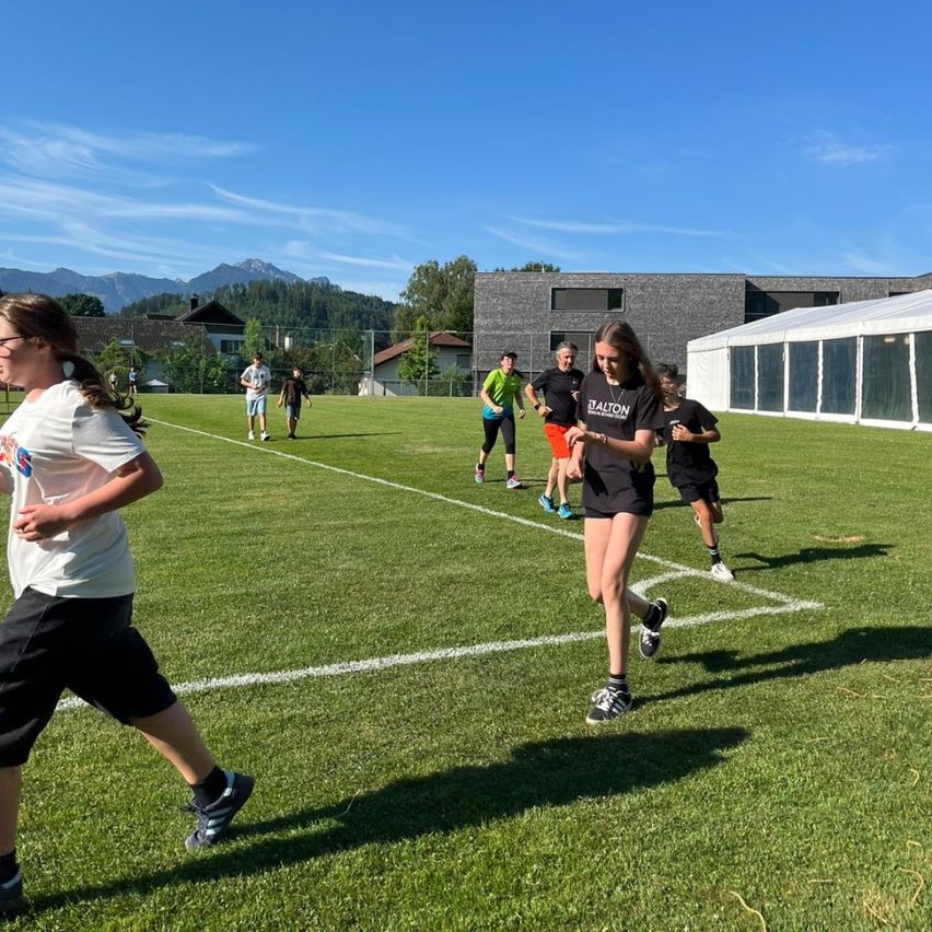 Menschen laufen auf einem Feld mit weißen Linien. Eine Frau in einem schwarzen Shirt läuft vorne. Hinter ihr laufen auch andere. Im Hintergrund sind Gebäude und Bäume zu sehen.