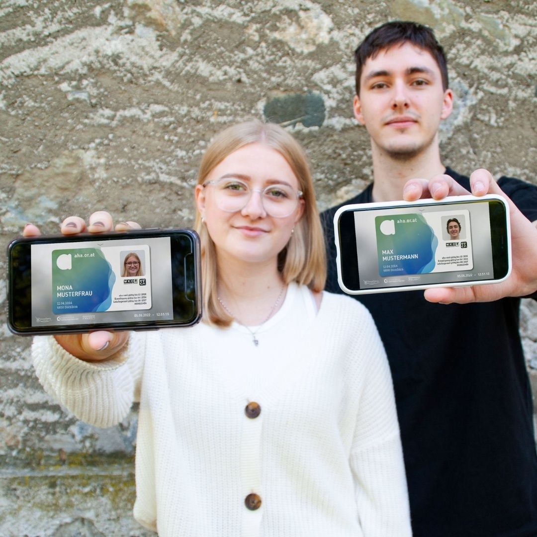 Eine Frau und ein Mann stehen vor einer Steinmauer, jeder hält ein Smartphone. Sie lächeln beide und zeigen ihre digitalen Ausweise auf den Bildschirmen. Die Frau trägt einen weißen Pullover und eine Brille. Der Mann ist in einem schwarzen T-Shirt.