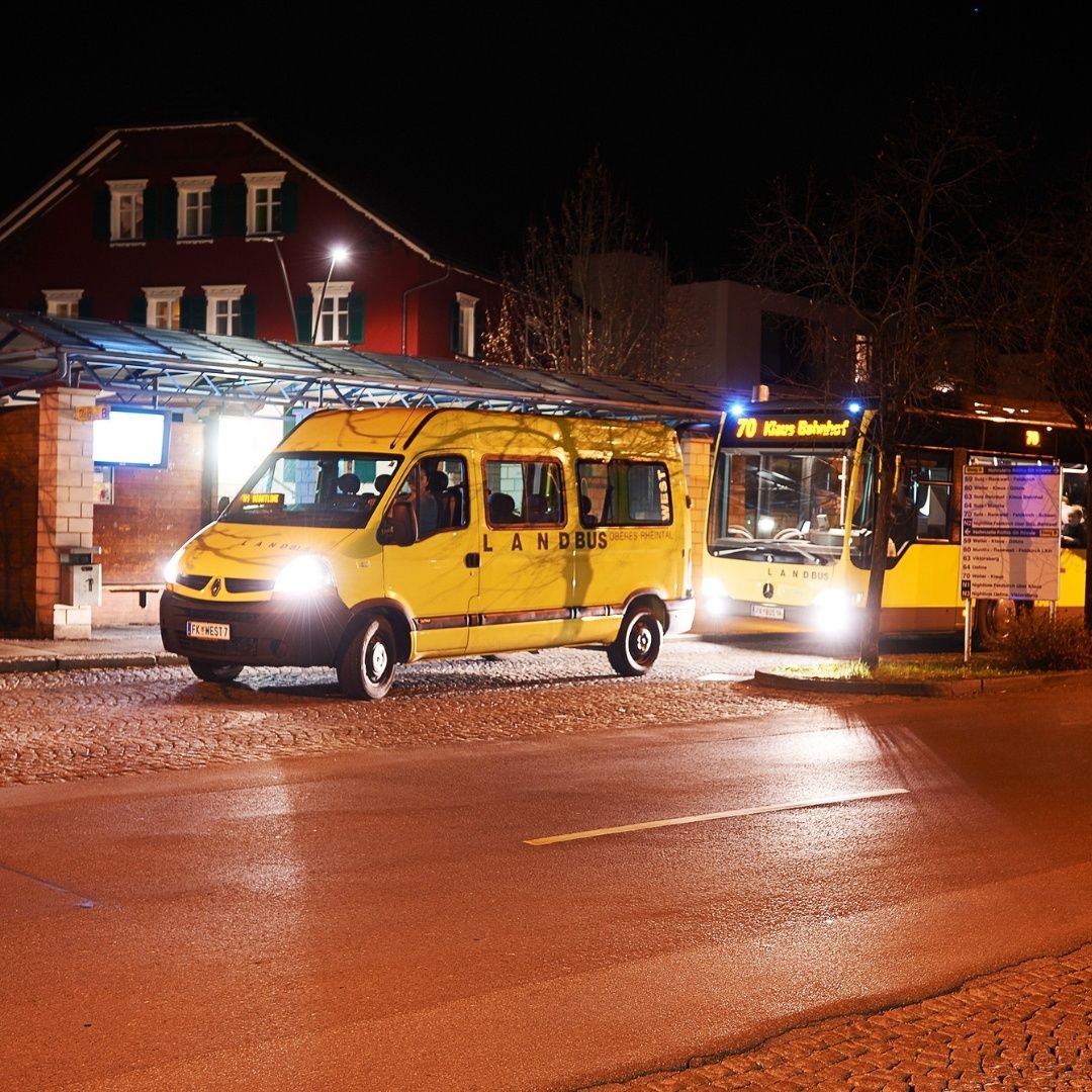 Ein gelber Bus mit der Aufschrift Landbus steht an einer Bushaltestelle. Ein weiterer Bus ist dahinter geparkt. Gebäude befinden sich auf der linken Seite.