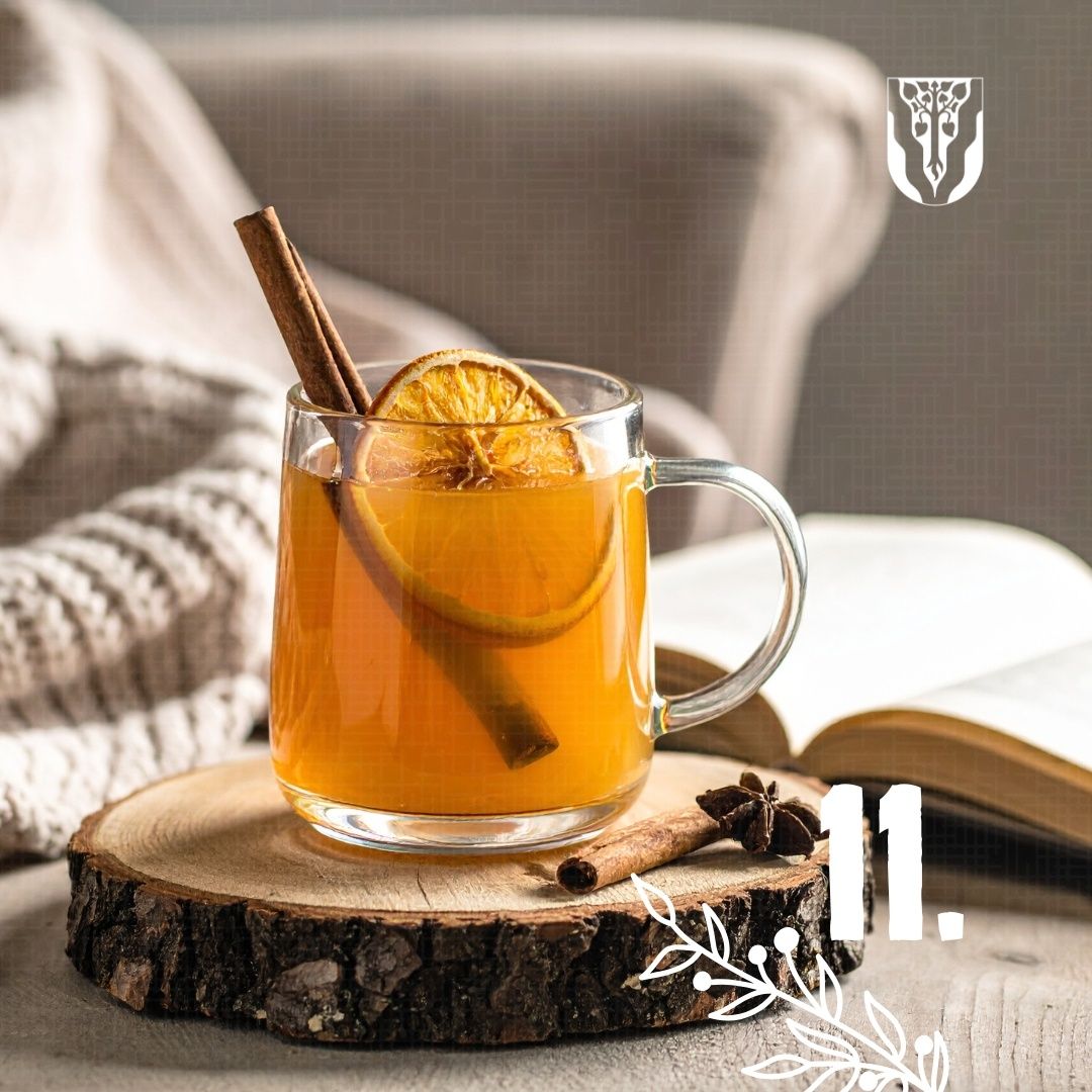 Ein warmes Getränk in einem Glasbecher mit Zimt und Orangenscheiben steht auf einer Holzoberfläche. Ein offenes Buch und ein gemütlicher Stuhl sind in der Nähe.