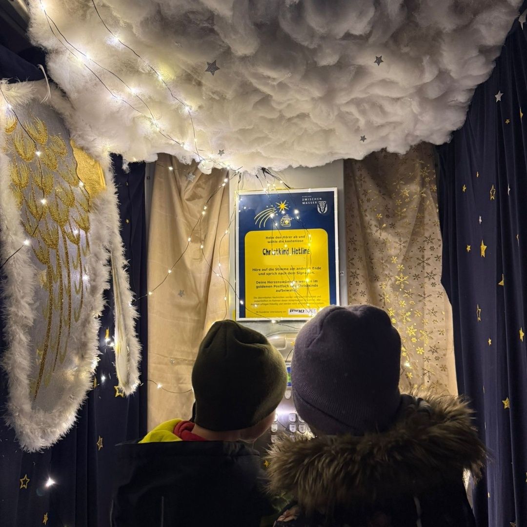 Zwei Kinder stehen in einer weihnachtlich dekorierten Kabine. Sie schauen nach oben zu einer sternübersäten Decke. Ein Poster an der Wand sagt 'Christkindl Hetling'.