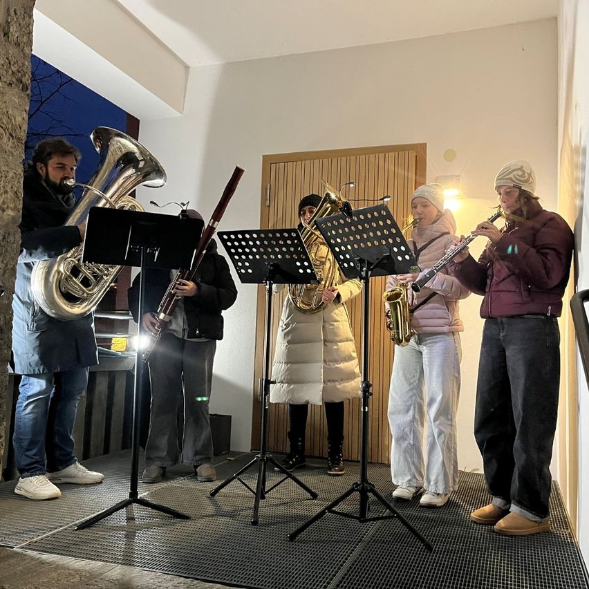 Fünf Personen stehen in einer Reihe und spielen Musikinstrumente, darunter Saxophon, Tuba und Flöte. Sie stehen vor einem Gebäude mit einer Holztür und einem Fenster. Der Boden ist mit einem schwarzen Teppich bedeckt.