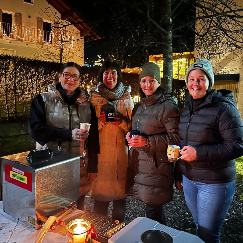 Vier Frauen stehen nachts draußen, lächeln und halten Tassen. Sie sind warm in Jacken und Mützen gekleidet. Eine Kaffeemaschine und eine Kerze stehen auf dem Tisch vor ihnen. Dahinter befindet sich ein Haus mit Lichtern und einem Baum.