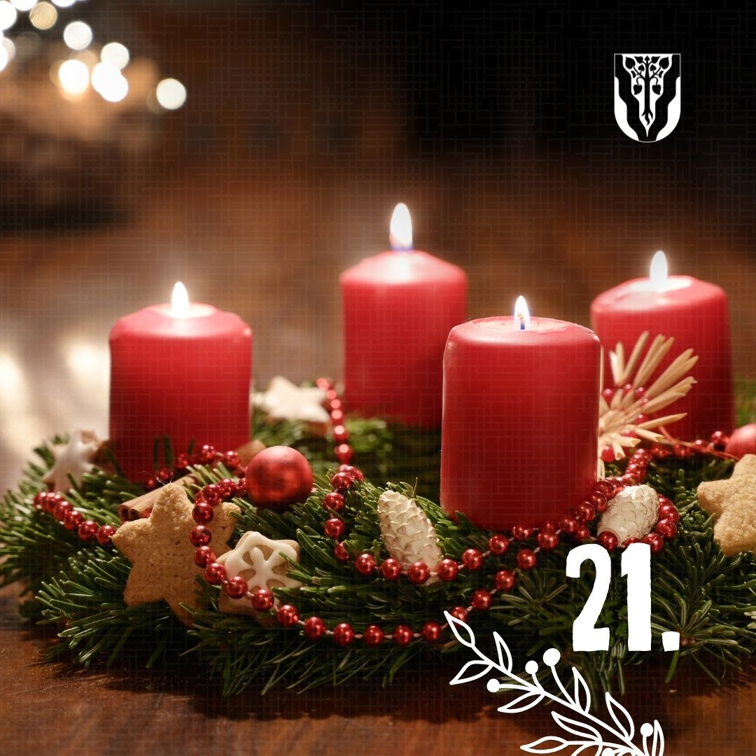 Eine Adventskrone mit vier roten Kerzen, einem Logo oben und der Zahl 21 in der Mitte. Die Krone ist mit Weihnachtsplätzchen, Tannenzapfen und roten Perlen geschmückt.