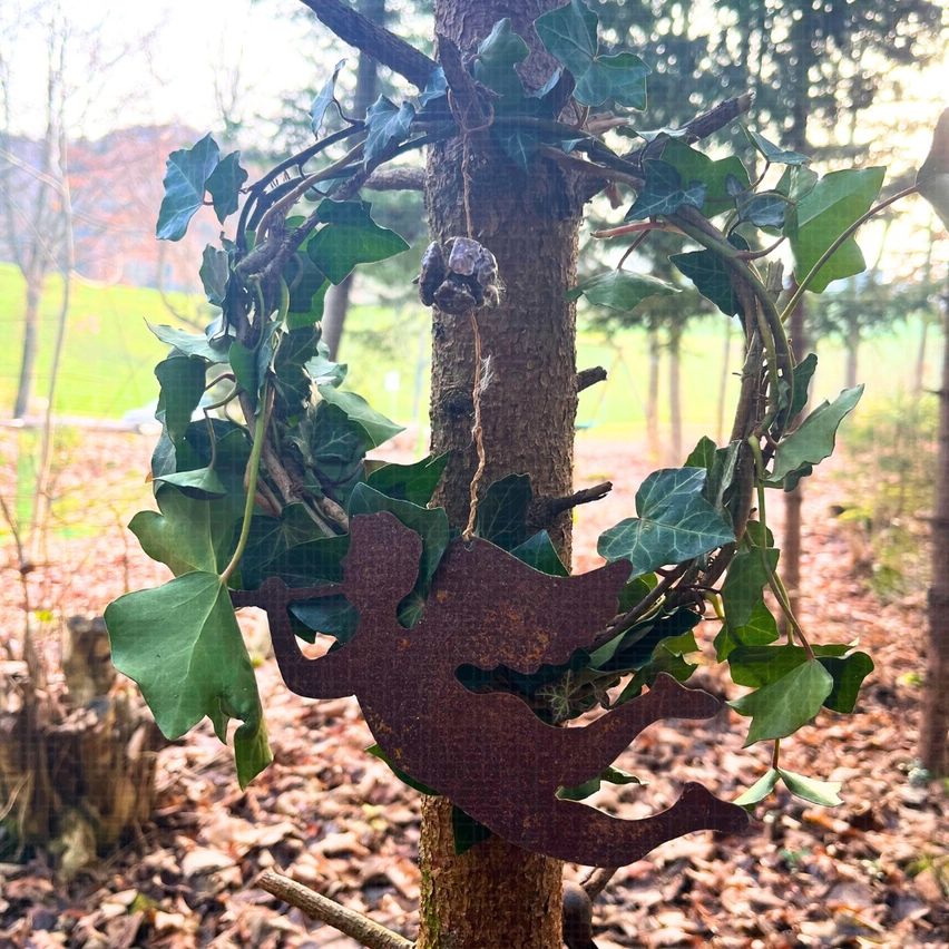 Ein Baum mit einem Kranz aus Efeublättern hängt an seinem Stamm und zeigt ein metallisches Engeldesign. Der Baum steht in einem Wald mit trockenen Blättern und einem Feld im Hintergrund.