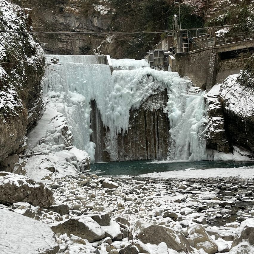 Eine verschneite Landschaft zeigt einen großen Wasserfall, der über Felsen fließt. Der Bereich ist schneebedeckt und oben befindet sich eine Holzbrücke.