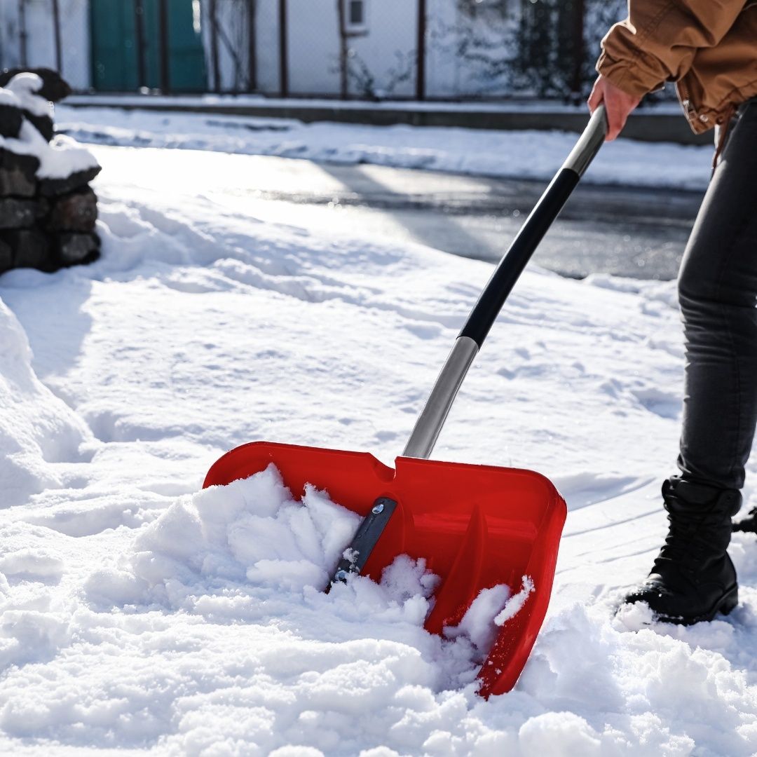 Eine Person benutzt eine rote Schneeschaufel auf einer verschneiten Straße. Sie trägt Stiefel und eine braune Jacke.