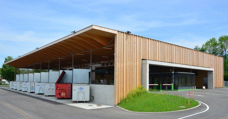 Ein modernes Gebäude mit Holzverkleidung und einem großen Vordach, mit mehreren Lagerboxen und einem grünen Pflanzenbereich davor.