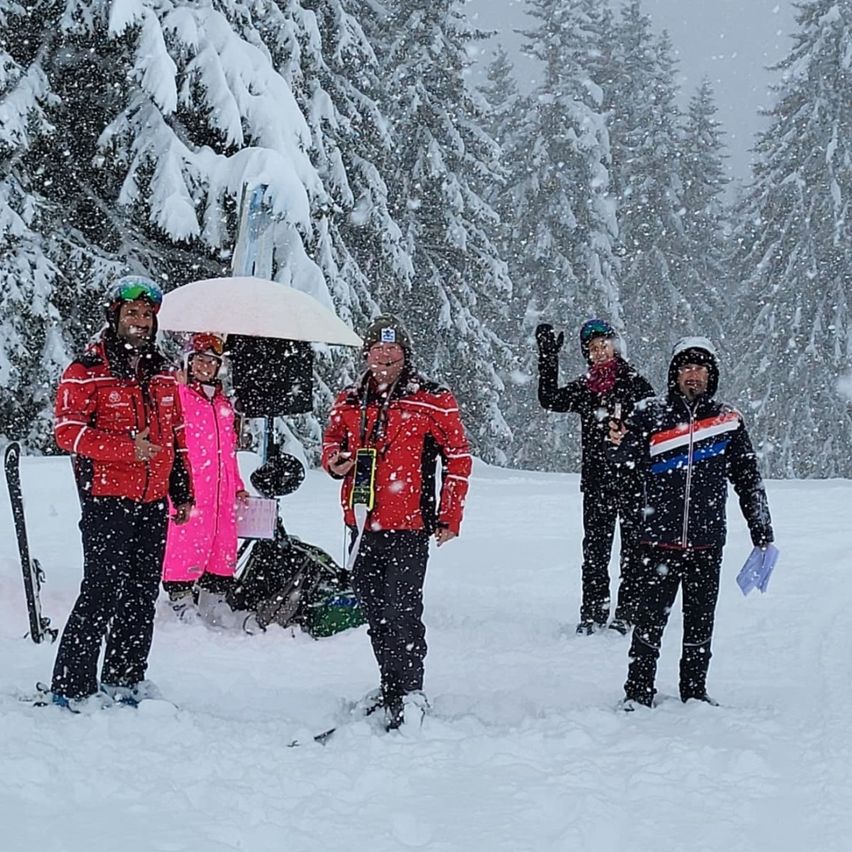 Fünf Personen, die Skijacken und Schneehosen tragen, stehen im Schnee. Sie lächeln und posieren für ein Foto. Der Mann auf der rechten Seite hält ein Papier.