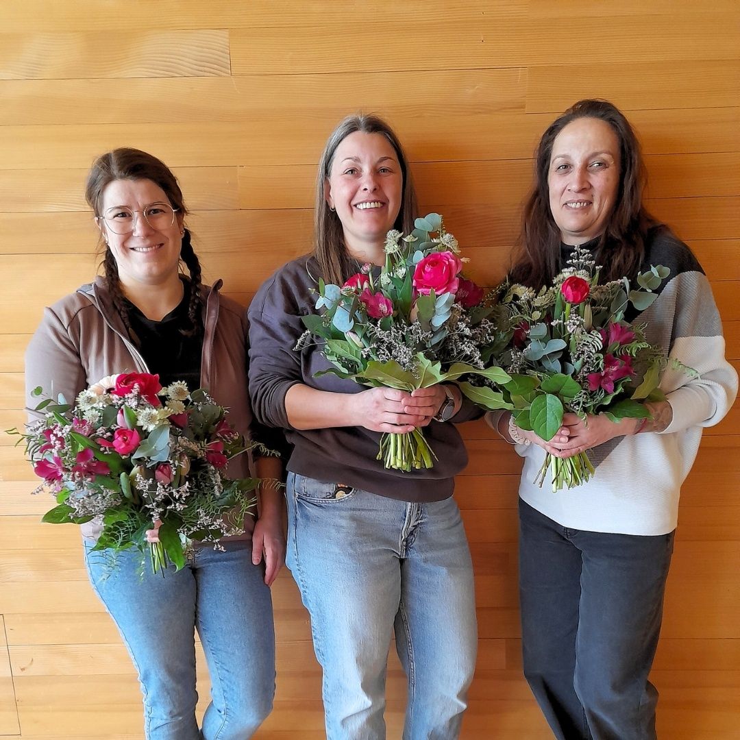 Drei Frauen stehen zusammen und halten jeweils einen Blumenstrauß in der Hand. Sie lächeln und posieren für ein Foto vor einer Holzwand.