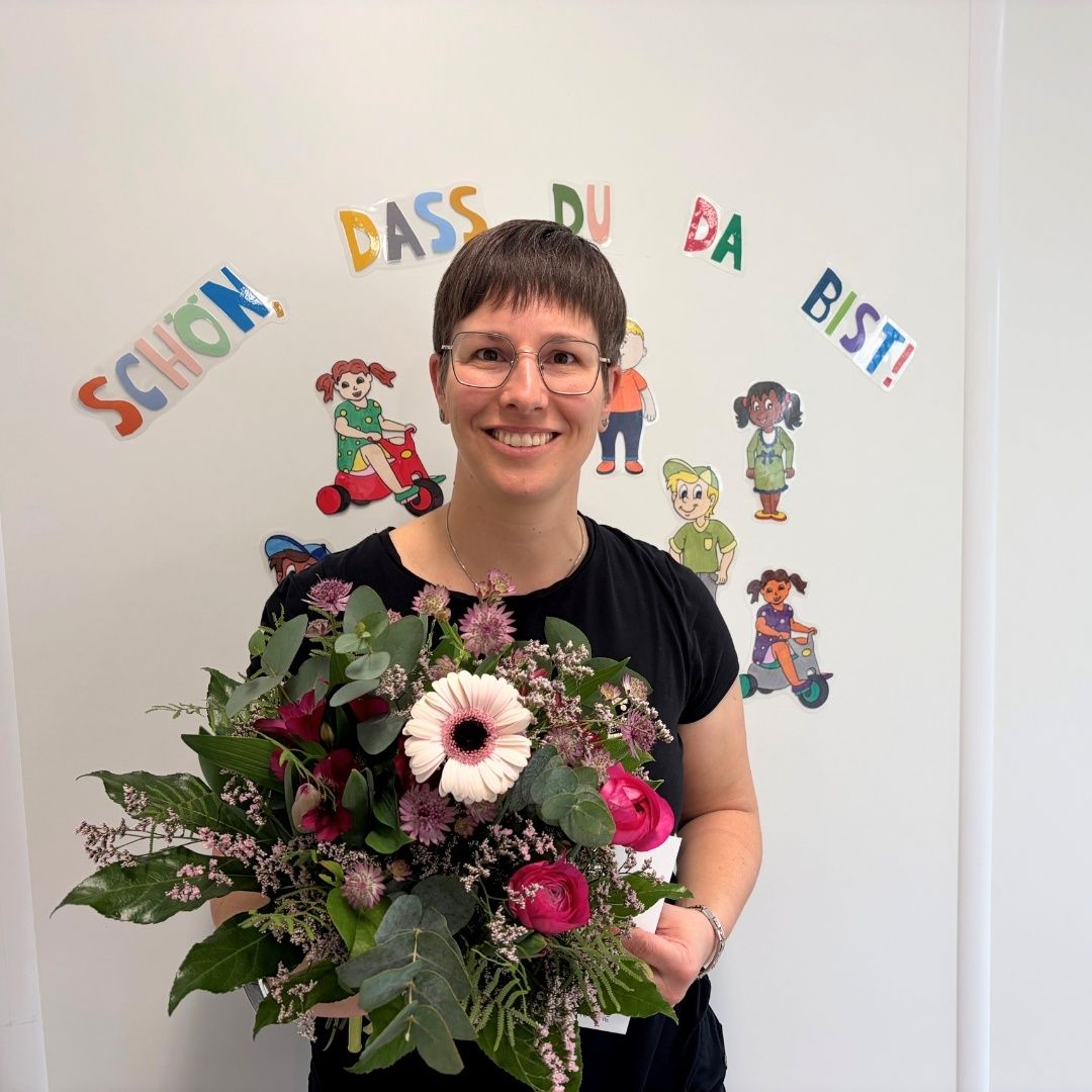 Eine Frau mit Brille hält einen Blumenstrauß in einem Raum mit einer weißen Wand, die mit bunten Aufklebern verziert ist. Die Aufkleber zeigen Kinder auf Rollern mit Texten auf Deutsch.